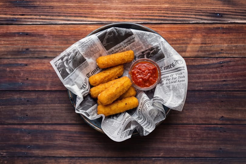 Golden fried mozzarella sticks and a small bowl of marinara sauce on newspaper in a basket.