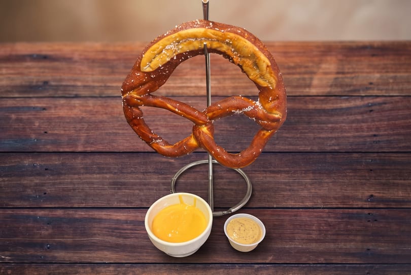 Large salted pretzel on a stand with cheese sauce and mustard on a wooden table.