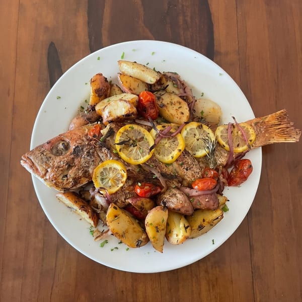 Whole roasted fish with lemon slices, roasted potatoes, cherry tomatoes, and red onion on a white plate.