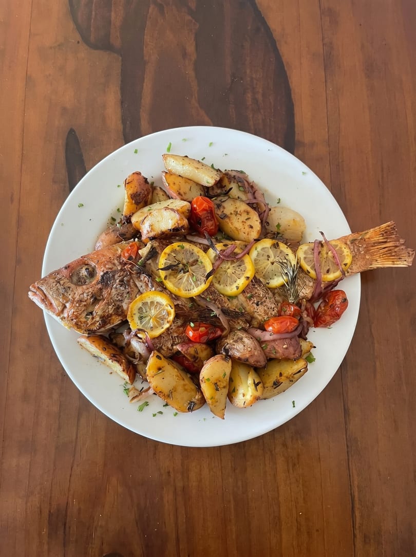 Whole roasted fish with lemon slices, roasted potatoes, cherry tomatoes, and red onion on a white plate.